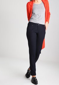 Tmavě modré kalhoty s upraveným střihem a jemnou texturou, doplněné šedým vzorovaným topem a jasně červeným kardiganem. Černé baleríny dokončují vzhled.