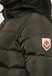 Mørkegrønne pufferjakke med quiltet mønster, brun lynlåstrim, justerbar hætte og en kremfarvet stofmærke med logo på skulderen.