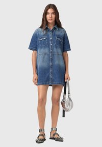 Robe chemise en denim avec des manches courtes, un devant boutonné, deux poches poitrine et un dégradé de couleur clair à foncé. Portée avec des sandales métalliques et un sac argenté.