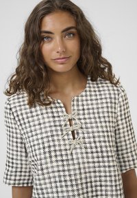 Košulja kratkih rukava u krem i crnoj kariranoj uzorku, s izrezom u obliku ključa te dvije dekorativne mašne od svijetlosive tkanine.