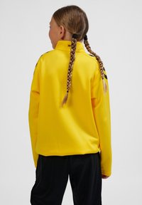 Gul långärmad jacka i slät tyg, med hög krage och svarta detaljer på axlarna. Kombinerad med svarta byxor.