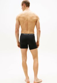 Boxer shorts de algodón negros con una cinturilla blanca de "TOMMY HILFIGER", diseño ajustado, textura suave, exhibidos en un modelo masculino visto desde atrás.