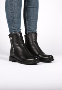 Botas de tobillo de cuero negro con un acabado texturizado, cierre de cremallera lateral, punta redondeada y tacón bajo. Combinadas con jeans azules.