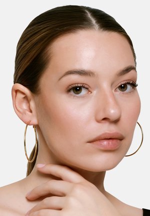 Donna dalla pelle chiara e capelli castani raccolti, che indossa grandi orecchini a cerchio dorati, toccandosi delicatamente il collo con la mano.