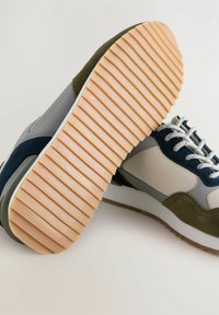 Tenisky s béžovou tkaninou, panely v námořnické a olivově zelené suede, bílé tkaničky a silnou hnědou gumovou podrážkou s hlubokými horizontálními drážkami.