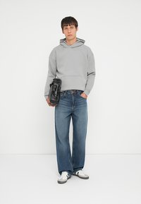 Grå hættetrøje med forlommer, iført løse blå denimjeans. Sort taske båret under armen, hvid og grå sneakers. Enkel baggrund.