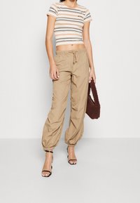 Lichtgewicht, loszittende broek in een beige kleur met elastische boorden, gecombineerd met een cropped top met meerdere strepen. Houdt een bruine handtas met een zachte, pluizige textuur vast.