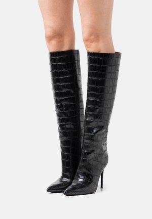 Zwarte leren laarzen met krokodillenpatroon tot aan de knie, met spits toelopende neuzen en naaldhakken, gedragen op blote benen.