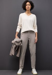 Kvinna med lockigt hår som står mot en mörk vägg, iförd en krämfärgad tröja, beiga byxor, vita sneakers och håller en taupe-färgad kappa i ena handen.