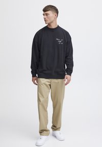 Black sweatshirt with white text "TELL IT LIKE IT IS," paired with beige trousers and white sneakers. Features a relaxed fit and crew neck.