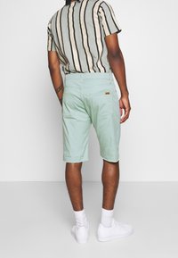 Man wearing light green knee-length shorts, a vertically striped beige and black t-shirt, white socks, and white sneakers, standing with back facing forward.