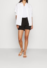 Chemise blanche boutonnée portée sur un débardeur blanc, associée à un short noir à cordon de serrage et des sandales marron clair à brides. Design simple.