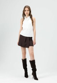 Witte mouwloze top met col, gecombineerd met een bruine minirok en zwarte kniehoge laarzen met hak. Soepele stof, modern silhouet.