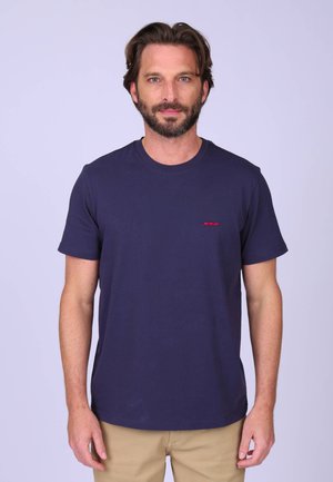 Homme barbu portant un t-shirt bleu marine à manches courtes et un pantalon beige, debout devant un fond clair uni.