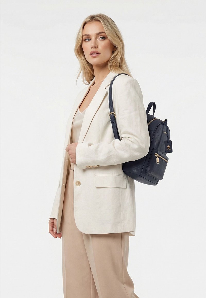 Femme blonde portant un blazer blanc et un pantalon beige porte un sac à dos en cuir bleu marine sur son épaule, regardant légèrement sur le côté.