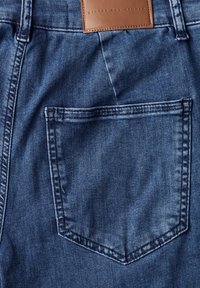 Tissu en denim bleu foncé avec un détail de poche arrière. Présente une étiquette en cuir avec l'inscription "STREET ONE STUDIO" et des coutures contrastées.