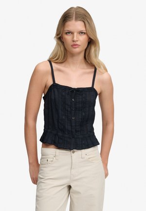 Kvinde med langt blondt hår iført en sort, knaplukket, ærmeløs top med flæsekant og lyse bukser mod en ensfarvet baggrund.