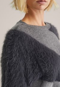 Grauer Pullover aus einem strukturierten, dunkelgrauen, fellähnlichen Material an einer Schulter, mit Rundhalsausschnitt und gerippten Strickdetails am Saum.
