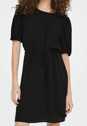 Woman wearing a knee-length black dress with short puff sleeves and a tied waist belt.