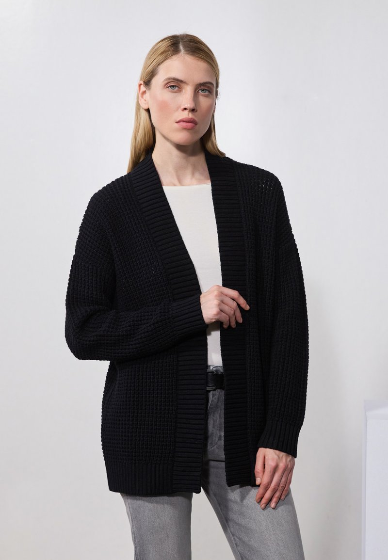 Schwarzer, gestrickter Cardigan mit offenem Vorderteil und langen Ärmeln, der strukturierte Muster und eine lockere Passform aufweist. Tragen über einem hellen Oberteil.