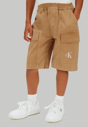 Kind trägt beige Cargo-Shorts mit Calvin Klein-Logo, weiße Socken und weiße Turnschuhe vor einem schlichten grauen Hintergrund.