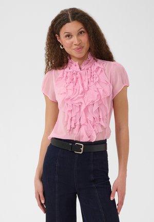 Kvinde med krøllet hår iført en gennemsigtig pink flæsebluse og mørke højtaljede bukser med sort bælte, stående mod en hvid baggrund.