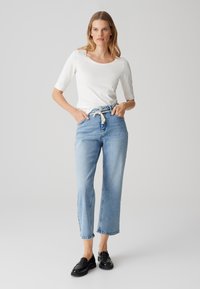 Un haut blanc ajusté à manches trois-quarts associé à un jean taille haute bleu clair avec une ceinture en tissu et des ourlets bruts. Des mocassins noirs complètent le look.