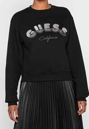 Personne portant un sweat-shirt noir avec un texte argenté "GUESS" et "California", associé à une jupe plissée noire.