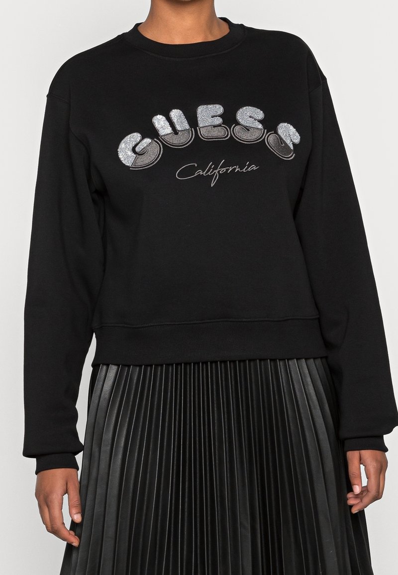 Personne portant un sweat-shirt noir avec un texte argenté "GUESS" et "California", associé à une jupe plissée noire.