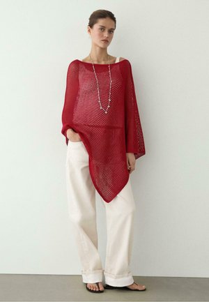 Femme debout contre un mur, portant un poncho rouge en maille ample, un pantalon blanc à jambes larges, un long collier et des sandales noires, une main dans la poche.