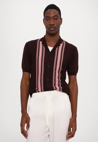 Cămașă tricotată maro cu mâneci scurte, cu dungi verticale roz și guler ascuțit. Butoane negre pe toată lungimea din față, purtată cu pantaloni albi.