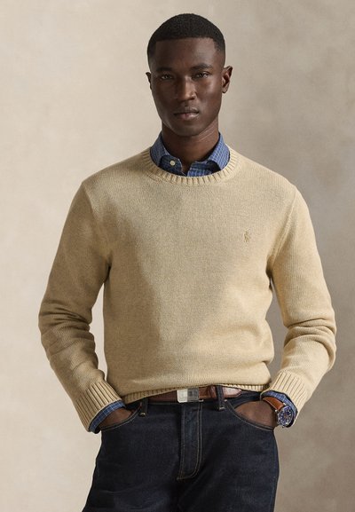 Beiger Strickpullover mit rundem Ausschnitt, darüber ein blau kariertes Hemd. Dunkle Jeans und ein brauner Gürtel vervollständigen den Look.