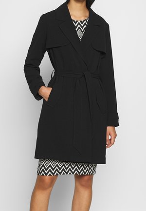 Woman wearing a black belted trench coat over a black and white zigzag patterned dress, standing with one hand in pocket.