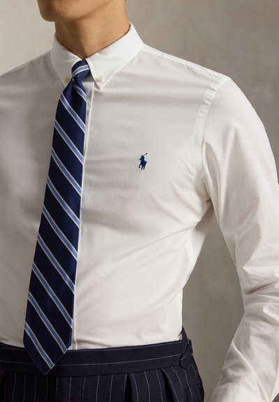 Man met een wit overhemd met knopen, een blauw gestreepte stropdas en donkere pantalon met fijne strepen, die een minimalistisch geborduurd logo toont.