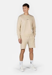 Felpa beige e pantaloni corti coordinati in tessuto morbido. La felpa presenta un collo rotondo e un logo discreto; i pantaloni corti hanno un cintura elasticizzata.