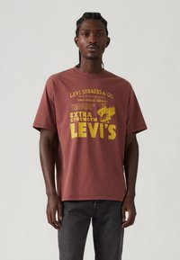 Bordeaux katoenen T-shirt met gele tekst en graphics, ronde crewneck, korte mouwen en een losse pasvorm. Gedrukte branding is zichtbaar.