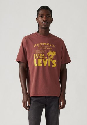 T-shirt en coton bordeaux avec texte et graphiques jaunes, col rond, manches courtes et coupe ample. La marque imprimée est visible.