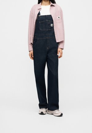 Person trägt dunkle Denim-Latzhose, weißes Hemd, rosa strukturierte Jacke und weiße Turnschuhe vor einfachem hellem Hintergrund.