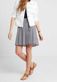 Vit denimjacka, marinblå topp, mönstrad utsvängd kjol i marinblått och vitt, bruna platta sandaler, stående pose med en ljus bakgrund.