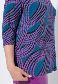 Blaues Oberteil mit einem Muster aus lila und schwarzen Punkten, mit 3/4-Ärmeln und einer lockeren Passform. Das Material wirkt weich und glatt.