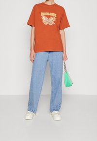 Orange bomulls t-shirt med fjärilsmotiv, kombinerad med ljusblå denim jeans med rak benpassform och en grön fluffig handväska. Vita sneakers bärs.