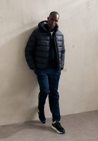Giacca a vento nera da uomo con cappuccio, pantaloni da jogging blu navy e sneakers nere con dettagli gialli. Tessuto morbido, design moderno, vestibilità rilassata.