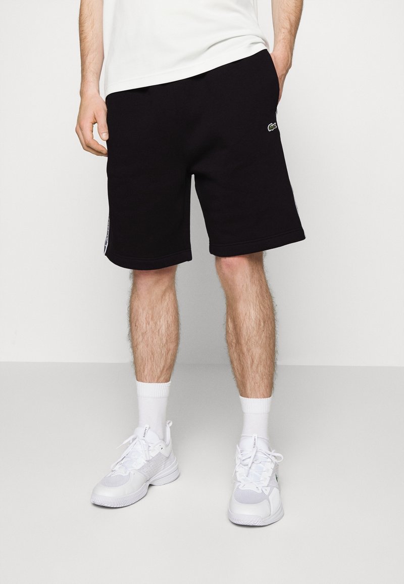 Svarta bomullsshorts med avslappnad passform, sidofickor och en vit logotyp på vänster ben. Matchas med vita sportskor och strumpor.