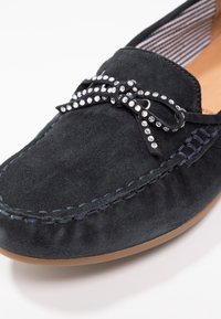 Chaussure en daim noire à enfiler avec un détail nœud orné de cristaux transparents, dotée d'une doublure intérieure rayée et de surpiqûres contrastées autour des bords.