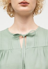 Κοντινό πλάνο ατόμου με σγουρά ξανθά μαλλιά που φοράει ανοιχτό πράσινο τοπ δεμένο με φιόγκο στο ντεκολτέ.