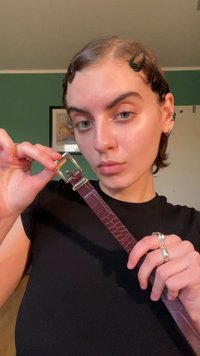 Burgundi bőr öv krokodilbőr textúrával és arany fém csattal, kézben tartva egy semleges háttér előtt.