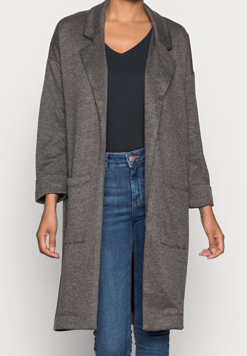 Manteau oversize gris texturé avec de larges revers porté sur un haut noir à col en V et un jean bleu taille haute.