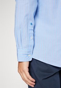 Blauw gestreept shirt met witte en rode lijnen, voorzien van een knoopmanchet en een klein geborduurd detail in blauw nabij de pols.