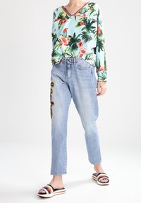 Top de manga larga azul claro con estampado floral tropical, combinado con jeans azul claro con bordado lateral. Sandalias deslizantes en blanco y negro.