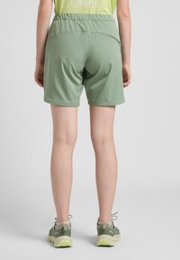 Pantaloni sportivi verdi con vita elastica, tasca con zip sul retro e texture liscia, abbinati a scarpe da trekking grigio-verdi.